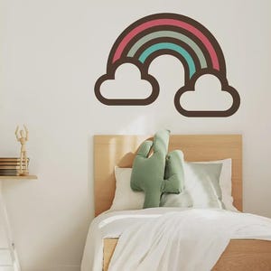 May include: A wall decal featuring a rainbow with thick dark brown outlines, a pink arch, and a teal arch, flanked by two white clouds. The design is displayed on a plain wall above a wooden bed frame with a green cactus-shaped pillow and white bedding.
