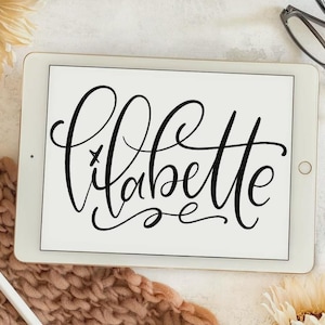 May include: A white tablet displaying the black cursive text lilabette in a stylized script font. The tablet is positioned alongside a white stylus, a pair of black-rimmed eyeglasses, a chunky knit peach-colored textile, and two cream-colored flowers.