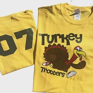 May include: Two yellow t-shirts. One shirt has the number "07" in dark green. The other shirt has a cartoon turkey with the words "Turkey Trotters" in black. The turkey is brown, red, and yellow.