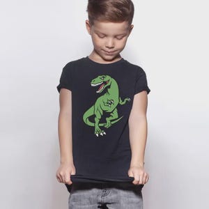 May include: A black short-sleeved t-shirt featuring a graphic print of a green Tyrannosaurus Rex dinosaur with white teeth and claws. The dinosaur is depicted in a standing position with its tail curved and mouth open, centered on the chest.