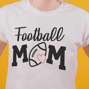 Puede incluir: Camiseta blanca con las palabras "Football MOM" en letras negras. Un gráfico de fútbol con un corazón está entre las palabras "MOM". Dos manos señalan la camiseta.