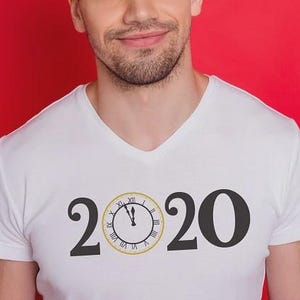 May include: White V-neck t-shirt with the year "2020" printed in black, featuring a clock design in the center. The clock shows the time as almost midnight. The background is a solid red.