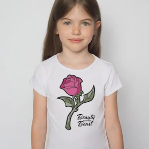 Op de afbeelding: Wit T-shirt met een roze roos, groene bladeren en stengel. De tekst "Beauty and the Beast" staat eronder. Het T-shirt wordt door een jong meisje gedragen.