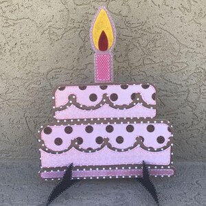 May include: A two-tiered birthday cake cutout featuring light pink icing with dark brown polka dots and scalloped brown trim. A single pink candle with a yellow and red flame sits on top. The cake is supported by two black metal stands.