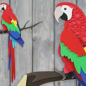 May include: Two layered paper craft parrots featuring vibrant red, green, yellow, and blue plumage with white and black facial details. The parrots are perched on brown branches. Text on the image includes SVG, DXF, PNG, and Craft with Sarah.