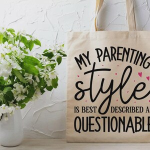 May include: A beige canvas tote bag featuring black text that reads MY PARENTING style IS BEST DESCRIBED AS QUESTIONABLE, accented with small pink dots and hearts. The bag has two fabric handles and is displayed in a vertical orientation.