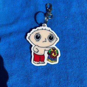 May include: A white keychain featuring a cartoon character with a large head, holding a colourful tie-dye shirt. The character has red shorts and a neutral expression. The keychain has a silver clasp and a key ring.