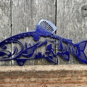 May include: A blue metal fish wall art piece with a mountain and Bigfoot silhouette design. The fish has a detailed fin and tail, and the design is cut out to reveal the background.