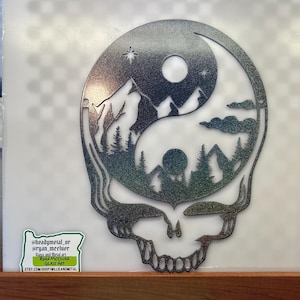 May include: Metal wall art featuring a yin-yang design within a skull outline. The yin-yang symbol depicts a mountain landscape with trees, clouds, and a star. The metal has a textured, iridescent finish. A sticker with social media handles and the words "Glass and Metal Art" is visible.