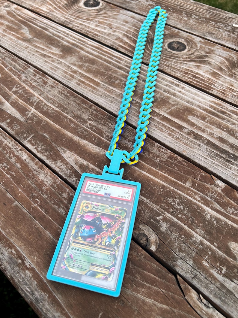 May include: A turquoise necklace with a matching card holder. The holder encases a Pokemon card with the text "M Venusaur EX". The chain is made of turquoise, yellow, and blue links. The card holder has the text "2016 Pokemon XY Evolutions".