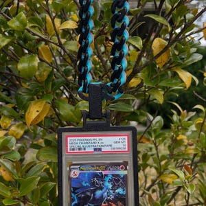 May include: A black and blue chain necklace with a pendant holding a Pokemon card. The card is encased in a clear plastic holder with text that reads "2025 POKEMON PFL EN MEGA CHARIZARD X ex SPECIAL ILLUSTRATION RARE". The chain is made of black and blue links.