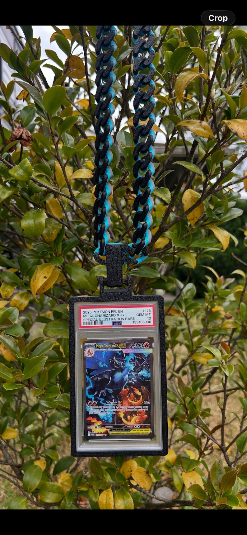 May include: A black and blue chain necklace with a pendant holding a Pokemon card. The card is encased in a clear plastic holder with text that reads "2025 POKEMON PFL EN MEGA CHARIZARD X ex SPECIAL ILLUSTRATION RARE". The chain is made of black and blue links.