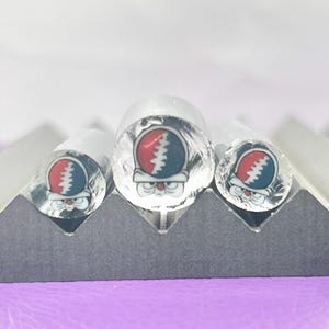 May include: Three clear, cylindrical objects with a red, white, and blue graphic of a skull with a lightning bolt on the front. The objects are arranged on a gray, angled surface, with a purple background.