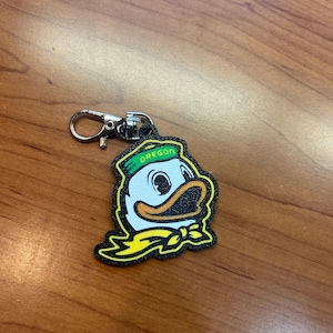 May include: A keychain featuring the Oregon Ducks mascot, a cartoon duck wearing a green hat with the word "OREGON" and a yellow bow tie. The keychain has a silver clasp.