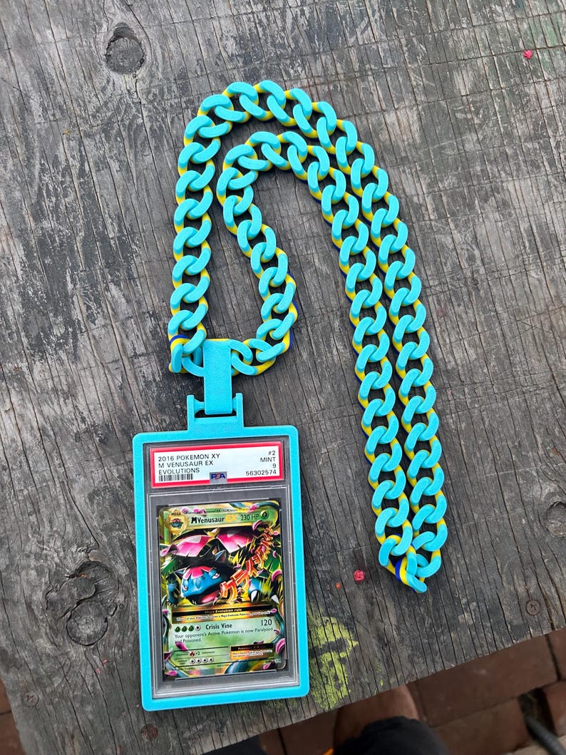 May include: A turquoise chain necklace with yellow accents, holding a graded Pok&eacute;mon card. The card is in a blue plastic case and features a colorful illustration of a Venusaur EX card from the 2016 Pok&eacute;mon XY Evolutions set.