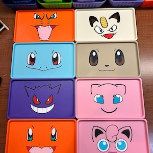 May include: Rectangular trays in various colors, each featuring a different cartoon character face. The trays are orange, blue, purple, pink, and beige. The faces have black outlines and are smiling or showing their teeth.