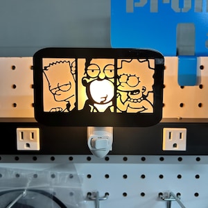 May include: Black night light featuring The Simpsons family: Bart, Homer, and Lisa. The lamp has a rectangular shape with the characters' silhouettes illuminated. The night light plugs into a standard electrical outlet.
