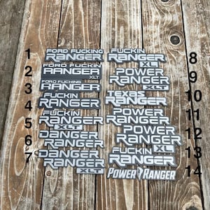 Può includere: Una collezione di stemmi per auto grigi e bianchi con varie frasi, tra cui "Ford Fucking Ranger" e "Power Ranger". Gli stemmi sono disposti su una superficie di legno invecchiato.