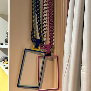 May include: Three colorful necklaces with matching rectangular frames hanging on a wall. The necklaces are blue, white, and pink, with coordinating frames in blue, pink, and white. The frames are empty.