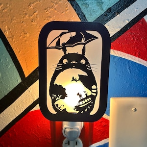 Puede incluir: Una luz de noche negra con la silueta de un personaje de dibujos animados bajo un paraguas. La luz de noche está enchufada a una toma de corriente blanca. El fondo es una pared colorida con pintura naranja, azul, roja y negra.