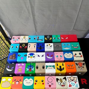 May include: A collection of colorful, hand-painted wooden boxes, each featuring a different cartoon character face. The boxes are arranged in neat rows on a black surface, with a variety of colors and expressions.