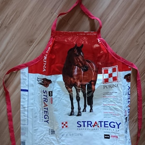 May include: An apron crafted from a repurposed Purina Strategy horse feed bag. The apron features a red and white color scheme with a central image of a brown horse. Text includes Purina, Strategy Professional Formula GX, and Net Wt. 50 lb.