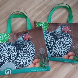 May include: Two tote bags with a brown background featuring a detailed illustration of a chicken with black and white feathers, a red comb, and a green handle. The bags have the text "NOW WITH 17% PROTEIN".