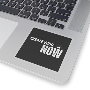 Può includere: Un adesivo quadrato nero con la scritta bianca "CREATE YOUR NOW". L'adesivo è posizionato sulla superficie argentata di un laptop, vicino alla tastiera. Il design include una grafica a raggi di sole.