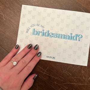 以下が含まれることがあります： 青色の「Will you be my bridesmaid?」というテキストが書かれた白いカード。カードは繰り返し模様で装飾されています。「07.25.26」の日付がカードに印刷されています。指輪をした手がカードの隣にあります。