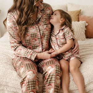 May include: Matching pajama sets featuring a repeating pattern of Santa Claus faces on a muted pink and olive green checkered background. The long-sleeved top and pants set, and the short-sleeved top and shorts set, both include dark red piping.