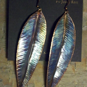 May include: A pair of handcrafted copper earrings in the shape of leaves. The earrings have a blue, green, and purple iridescent finish. The earrings are displayed on a black card with the text "HANDCRAFTED COPPER ORIGINALS BY JOHN ROSE".