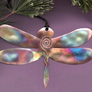 May include: A copper dragonfly ornament with iridescent colors and a spiral design in the center. The dragonfly is hanging from a black cord.