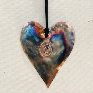 May include: A copper heart-shaped ornament with a spiral design in the center. The ornament is hanging from a black cord.