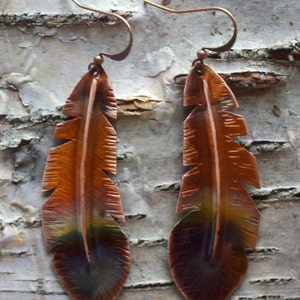 May include: A pair of copper feather earrings. The earrings are a warm copper color with a darker gradient at the bottom. The feathers have a detailed, textured design and are suspended from copper ear wires.