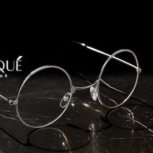 May include: Silver-rimmed eyeglasses with round lenses, resting on a dark marble surface. The glasses have a minimalist design with thin frames and temples. The brand name "ERATIQUE EYEWEAR" is displayed in white text.