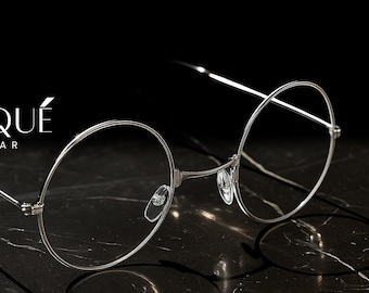 May include: Silver-rimmed eyeglasses with round lenses, resting on a dark marble surface. The glasses have a minimalist design with thin frames and temples. The brand name "ERATIQUE EYEWEAR" is displayed in white text.