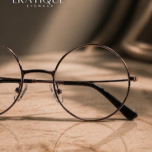 May include: Close-up of round, wire-rimmed eyeglasses with a brown metal frame and black temples. The glasses are set against a neutral background, with the brand name "ERATIQUE EYEWEAR" visible at the top.