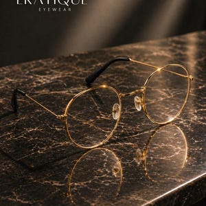 May include: Gold-rimmed eyeglasses with round lenses and black temples rest on a dark, marble-like surface. The brand name "ERATIQUE" and the word "EYEWEAR" are displayed in white text at the top. The glasses reflect on the surface.