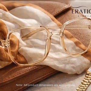 May include: Clear square-framed eyeglasses with a gold watch and a brown leather case. The glasses are resting on a cream and brown scarf. The watch has a gold band and a square face. The brand name "ERATIQUE EYEWEAR" is visible.