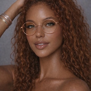 May include: A woman with curly auburn hair wearing round, gold-rimmed eyeglasses. The glasses have a thin frame and are positioned on the bridge of the nose. The image includes the text "ERATIQUE EYEWEAR".