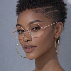 May include: A woman wearing round, rose-gold eyeglasses and large gold hoop earrings. The glasses have thin frames and clear lenses. The image includes the text "ERATIQUE EYEWEAR" at the top. The woman has short, curly hair and is looking at the camera.