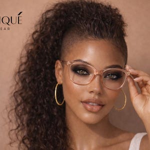 May include: A woman wearing clear-framed eyeglasses with a square shape. The glasses are held up with one hand. The model is also wearing gold hoop earrings. The brand name "ERATIQUE EYEWEAR" is visible in the upper left corner.