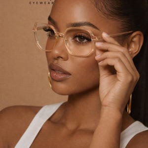 May include: Clear, square-framed eyeglasses with a light brown tint. The glasses are being held up by a person. The brand name "ERATIQUE EYEWEAR" is visible in the top left corner. The background is a solid, light brown color.