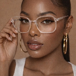 May include: Clear, octagonal eyeglasses with a transparent frame. The image features the brand name "ERATIQUE EYEWEAR" at the top. The model is wearing gold hoop earrings and a white tank top.