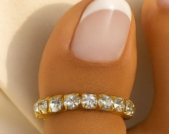May include: A gold-toned toe ring featuring a row of seven clear, round-cut prong-set crystals. The band is circular and metallic, designed to fit around a toe. The text ERATIQUE is visible in the upper right corner of the image.