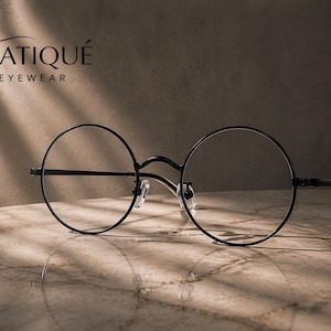 May include: Black round-frame eyeglasses with clear lenses. The glasses have a thin, minimalist design and are set against a marble surface. The brand name "ERATIQUE EYEWEAR" is displayed in the upper left corner.