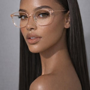 May include: Round, rose-gold eyeglasses with clear lenses. The glasses are worn by a person with long, dark hair. The brand name "ERATIQUE EYEWEAR" is printed at the top of the image.