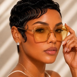 May include: Square-shaped eyeglasses with translucent amber-toned frames and yellow-tinted lenses. The upper right corner features the text ERATIQUE EYEWEAR in a black serif font. The design includes thin temples and a bridge.