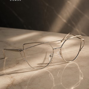 May include: Silver-toned eyeglasses with a unique cat-eye design. The frames are thin and delicate, with a modern aesthetic. The brand name "Ératiqué Eyewear" is displayed in the upper left corner. The glasses are set on a marble surface.