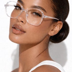 May include: Clear, square-framed eyeglasses. The glasses have a transparent frame and are worn by a person. The brand name "ERATIQUE EYEWEAR" is visible in the upper left corner. The person is wearing a white tank top.